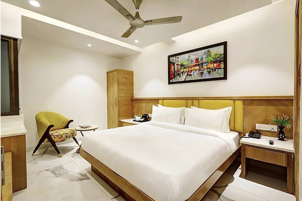 A modern hotel room with a large bed, a wooden headboard, a yellow armchair, a small table, a nightstand, a ceiling fan, and a painting on the wall.