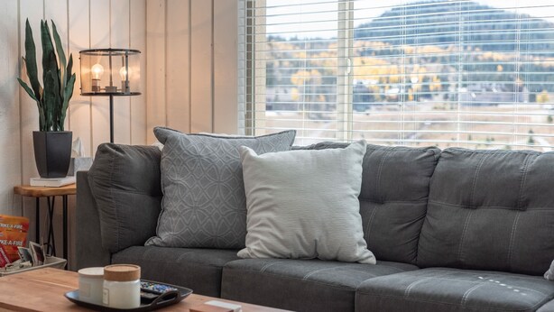 Un salon moderne avec un canapé d’angle gris, une petite table d’appoint avec une lampe, et une vue sur un paysage montagneux grâce à de grandes fenêtres.