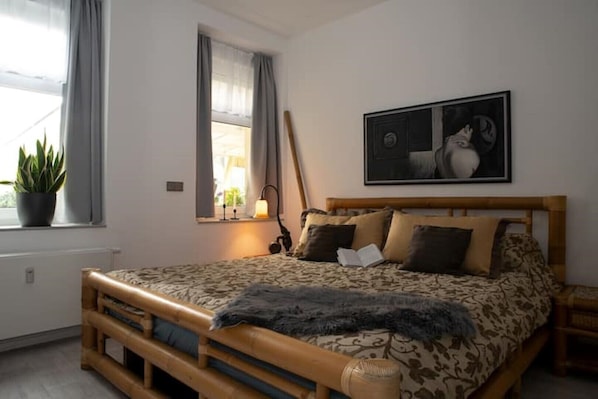 A bedroom with a wooden bed, a window with curtains, a potted plant, and a wall-mounted lamp.
