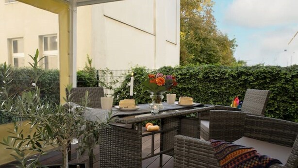 An outdoor patio with wicker furniture, a table, and a candle. There are plants and a hedge in the background. - NEU Luxus-apartment! Wasserbett-terrasse-smarttv (Magdeburg)