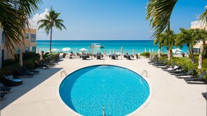 A pool area with lounge chairs and umbrellas, a clear view of the ocean, and a building with balconies. - Summer on Seven Mile Beach | Ocean View 2BR at Regal Beach Club (Seven Mile Beach)