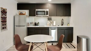 Una cocina compacta con armarios oscuros, una encimera blanca, microondas, lavavajillas y una pequeña mesa de comedor con dos sillas.
