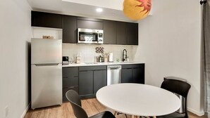 A modern kitchen with dark cabinets, a stainless steel refrigerator, a microwave, and a dishwasher. There is a round white table with black chairs in the center. - Moon's Studio apartment with Pool (Tempe)
