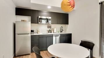 A modern kitchen with dark cabinets, a stainless steel refrigerator, a microwave, and a dishwasher. There is a round white table with black chairs in the center.