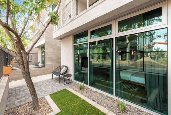 A modern building with large glass windows, a small patio with a tree and seating, and a view of a playground.