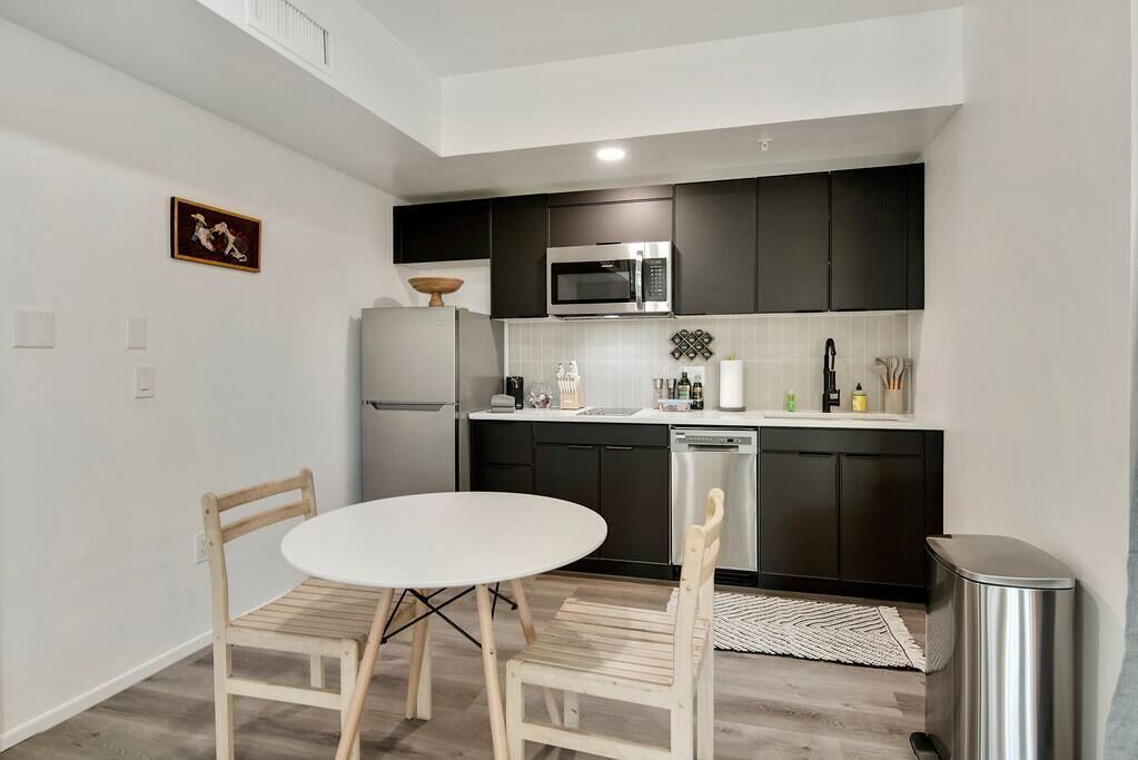 Eine kompakte Küche mit dunklen Schränken, einer weißen Arbeitsplatte, einer Mikrowelle, einem Kühlschrank und einer kleinen Essecke mit einem weißen Tisch und Holzstühlen.