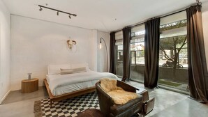 Ein modernes Schlafzimmer mit einem großen Bett, einem Ledersessel, einem karierten Teppich und Blick ins Freie durch große Fenster.