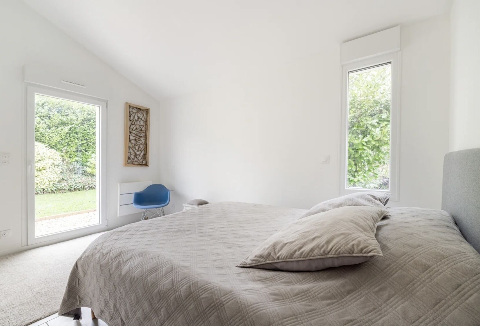 A bedroom with a large bed, a blue chair, and two windows.