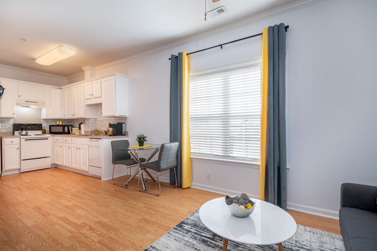 A modern kitchen with white cabinets, a dining area with a table and chairs, and a large window with yellow curtains.