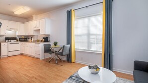 A modern kitchen with white cabinets, a dining area with a table and chairs, and a large window with yellow curtains. - Sunny Daze · Sunny Daze- Chic condo close to NASCAR (Concord)