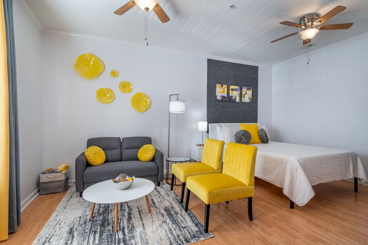 Una habitación de hotel moderna con un sofá gris, sillones amarillos, una cama blanca y un ventilador de techo.