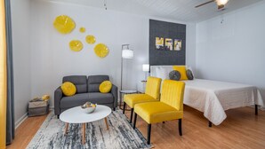 A modern hotel room with a gray sofa, yellow armchairs, a white bed, and a ceiling fan. - Sunny Daze · Sunny Daze- Chic condo close to NASCAR (Concord)