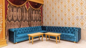 Une pièce avec un canapé en velours bleu, deux tables en bois, des rideaux décoratifs et un papier peint à motifs.