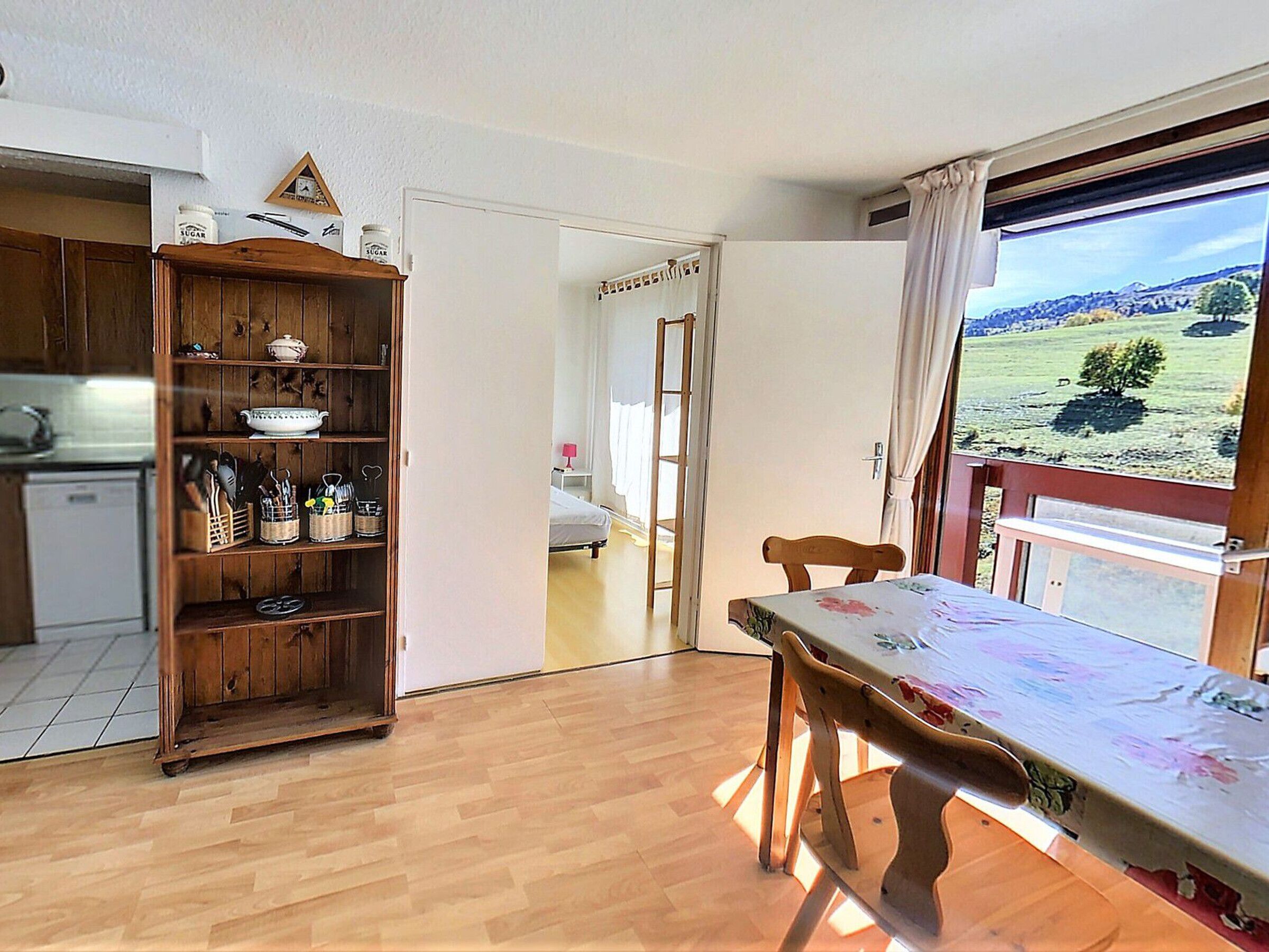 Ein Zimmer mit Esstisch aus Holz, Stühlen, einer Küchenzeile mit Holdekor und Blick auf eine grüne Landschaft durch ein Fenster.
