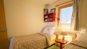 Um quarto pequeno e acolhedor com duas camas de solteiro, uma mesa de cabeceira com luminária, uma janela com cortinas e uma estante com livros e um ursinho de pelúcia.
