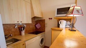 Cozinha compacta com armários de madeira, máquina de lavar louça, máquina de lavar roupa, micro-ondas e um pequeno balcão com chaleira e luminária.