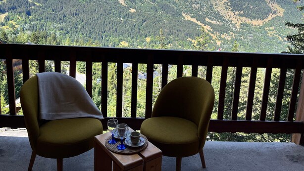 Varanda com duas cadeiras e uma mesa, com uma taça de vinho e uma chávena, com vista para uma paisagem montanhosa.