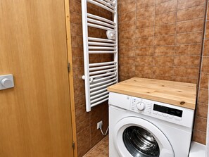 A compact laundry room with a front-loading washing machine, a wooden drying rack, and a tiled wall.