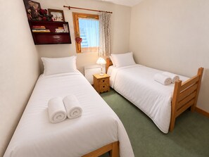 A twin room with two single beds, a bedside table with a lamp, a window with curtains, and a bookshelf with books and a teddy bear.