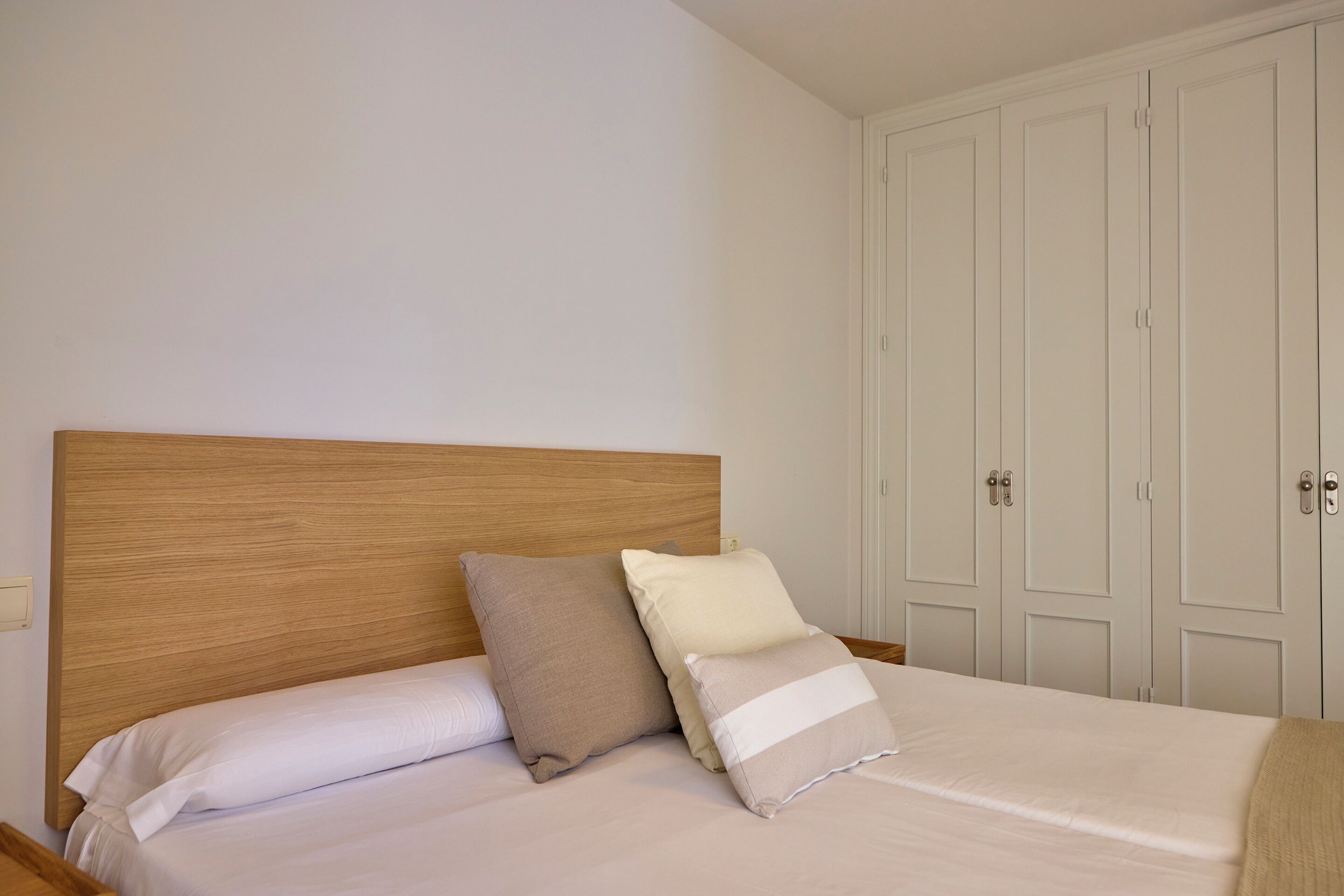 A bed with a wooden headboard, two pillows, and a white bedspread in a room with white walls and a white wardrobe.