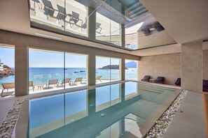 A modern infinity pool with clear views of the sea and boats, surrounded by lounge chairs and a high-ceilinged interior. - Universal Aquamarin Beach Houses (Andraitx)