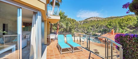 A balcony with two blue lounge chairs, a glass door leading to a room, and a view of a hilly landscape.