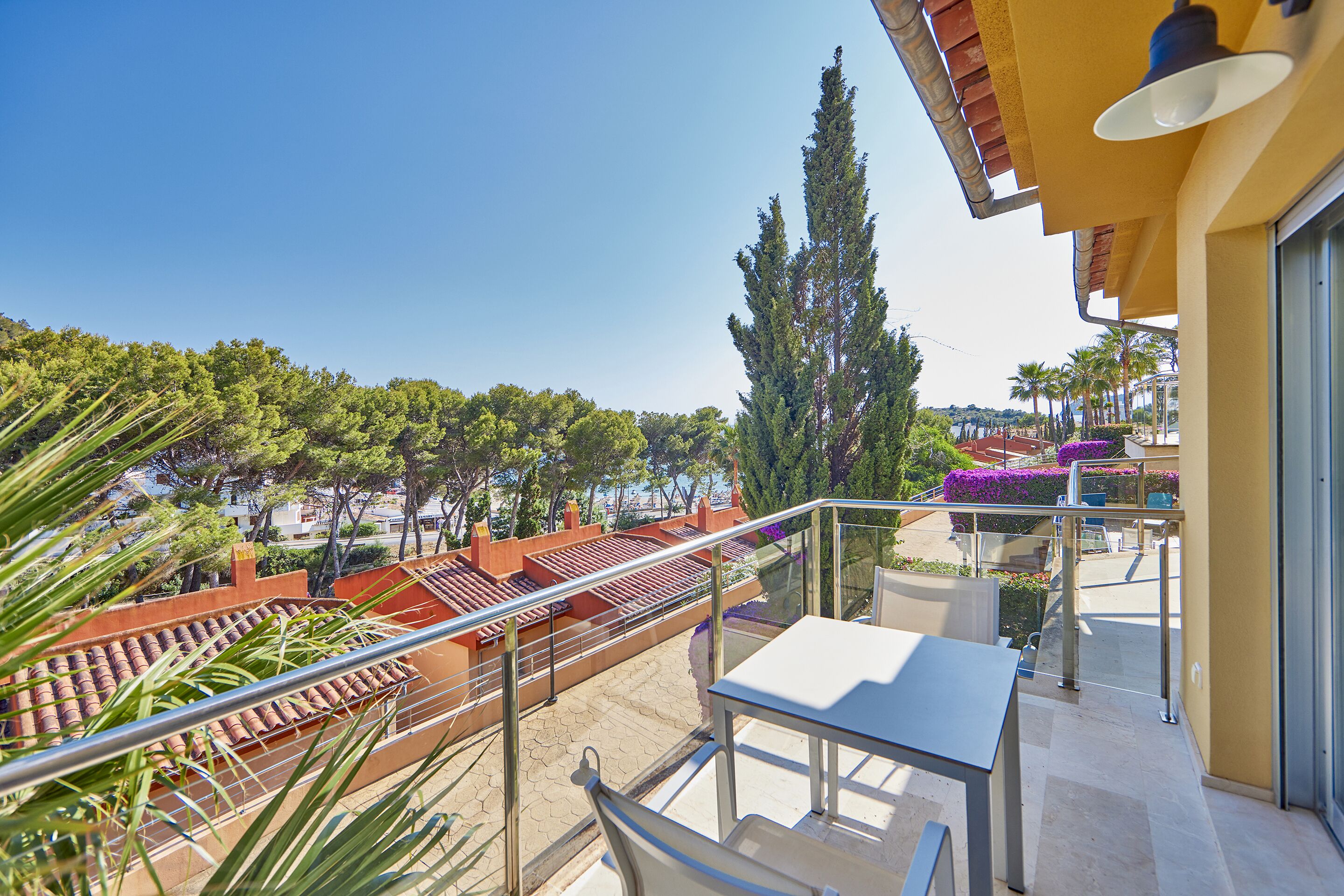 A balcony with a table and chairs, a railing, and a view of a landscaped area with trees and a building in the background.