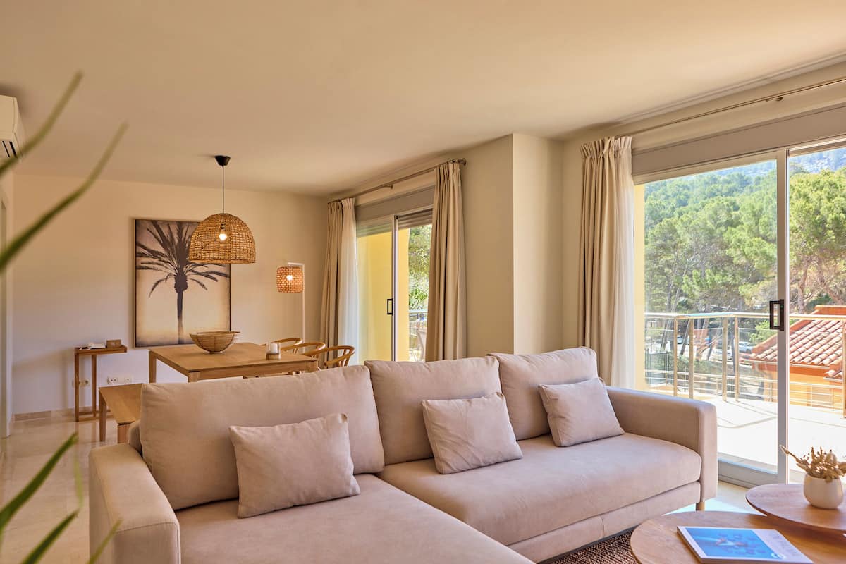 A modern living room with a beige sofa, a dining table, and a large window with a view of greenery.