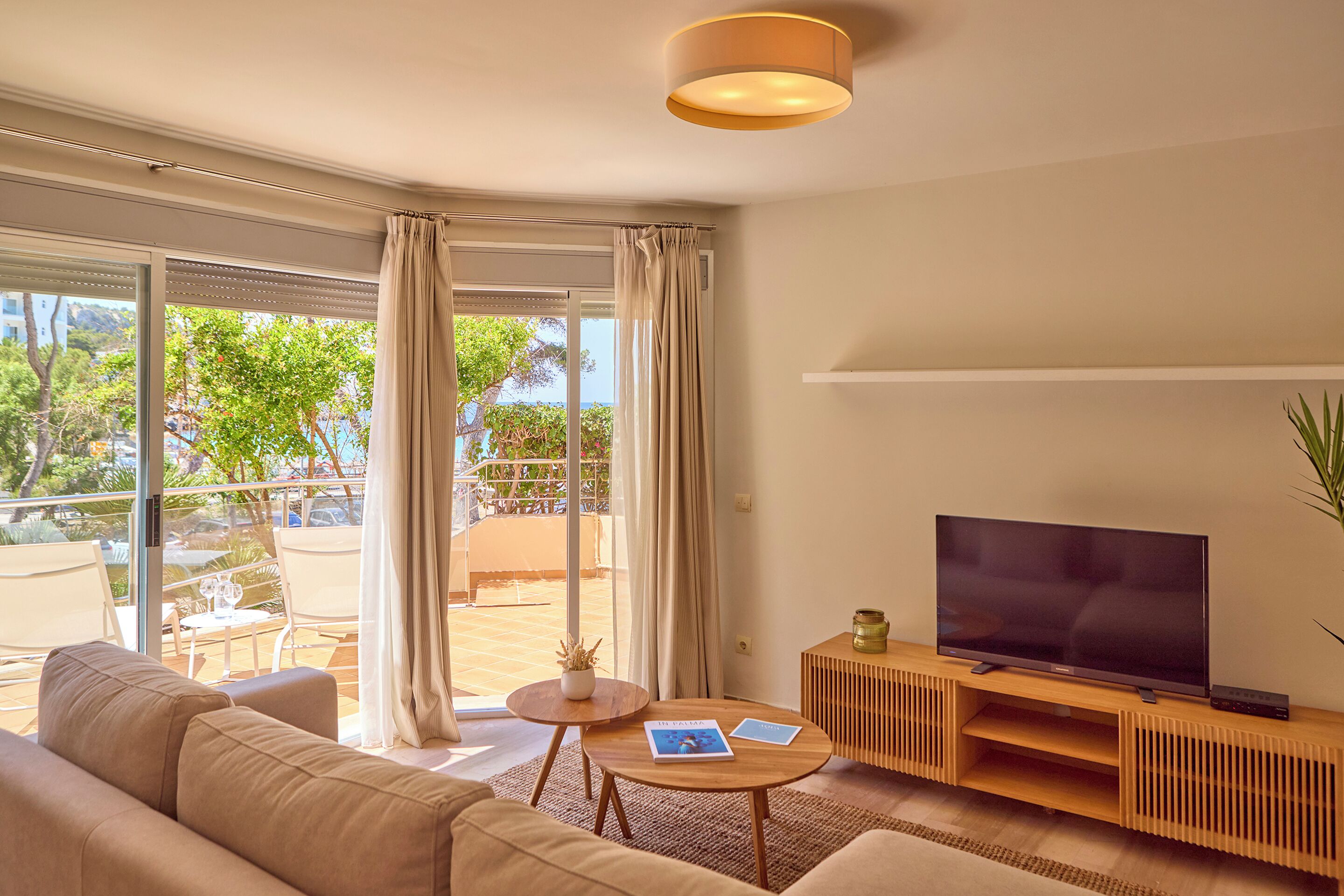 Ein modernes Wohnzimmer mit einer Couch, einem Couchtisch, einem Fernseher auf einem Holzgestell und Blick ins Freie durch große Fenster.
