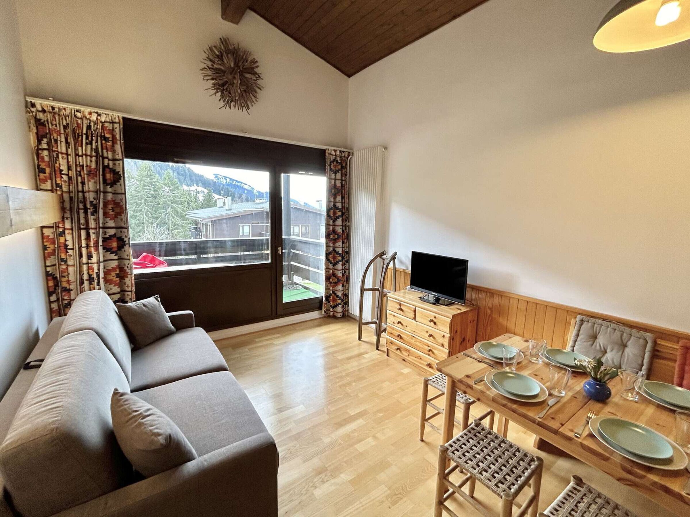 Un soggiorno con un divano, un tavolo da pranzo in legno apparecchiato per due persone, una televisione su un mobile in legno e vista su un edificio attraverso un'ampia finestra.