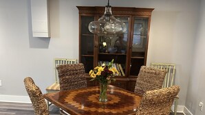Un comedor con una mesa de madera, sillas de mimbre, un florero con flores y un aparador al fondo.