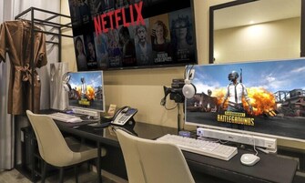 Una habitación con dos escritorios para computadora, cada uno con monitor, teclado y ratón. Hay un televisor de pared que muestra carteles de películas.