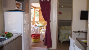 A compact kitchen with a refrigerator, a dining table with chairs, and a bed with a patterned bedspread. - Camping Tikayan Les Palmiers (Hyères)