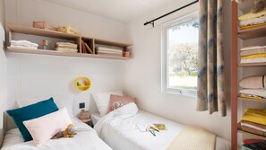 A bedroom with two beds, a window with curtains, a shelf with folded towels, and a small round yellow shelf with items. - Camping Tikayan Les Palmiers (Hyères)
