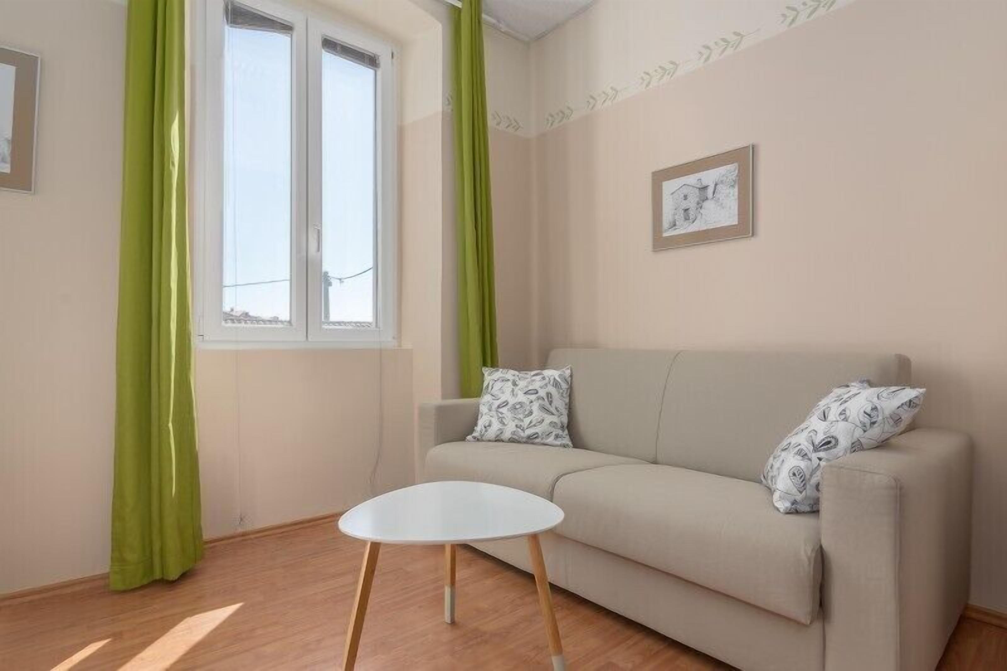 A living room with a beige sofa, two patterned pillows, a small white table, and a window with green curtains.