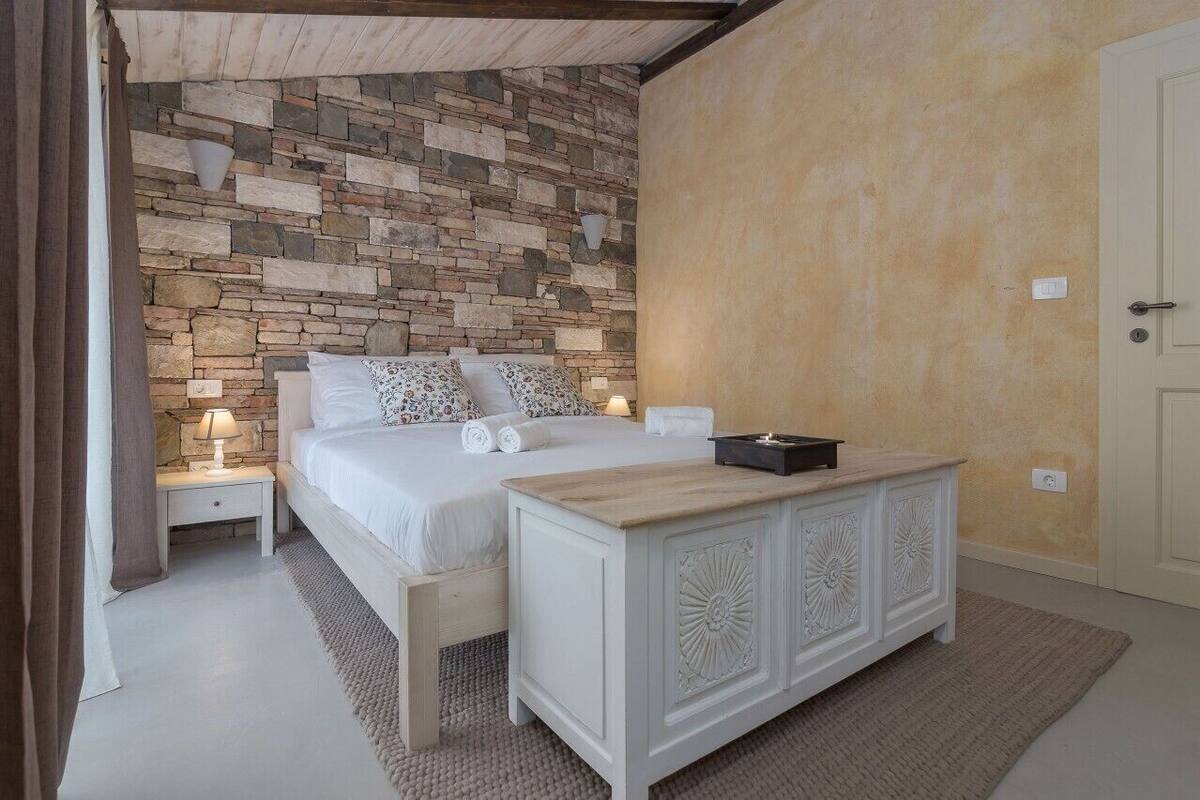 A bedroom with a stone wall, a bed with white linens, a bedside table with a lamp, and a wooden nightstand.