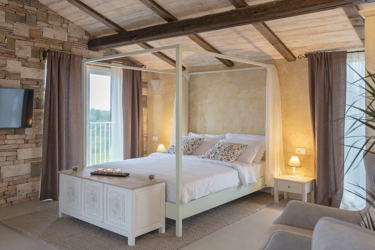 A four-poster bed with a white frame, a stone wall, a flat-screen TV, and a nightstand with a lamp.