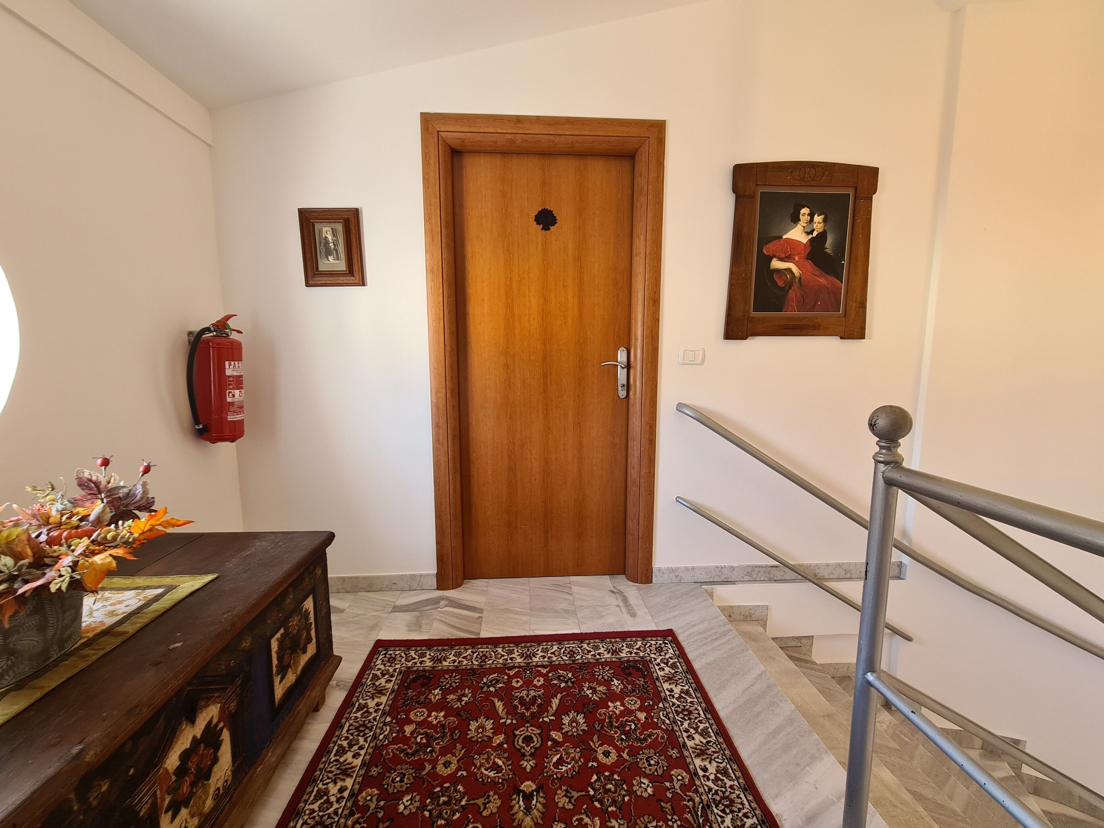 Un couloir avec une porte en bois, un extincteur, un tableau encadré, une petite table avec un vase et un tapis rouge à motifs.