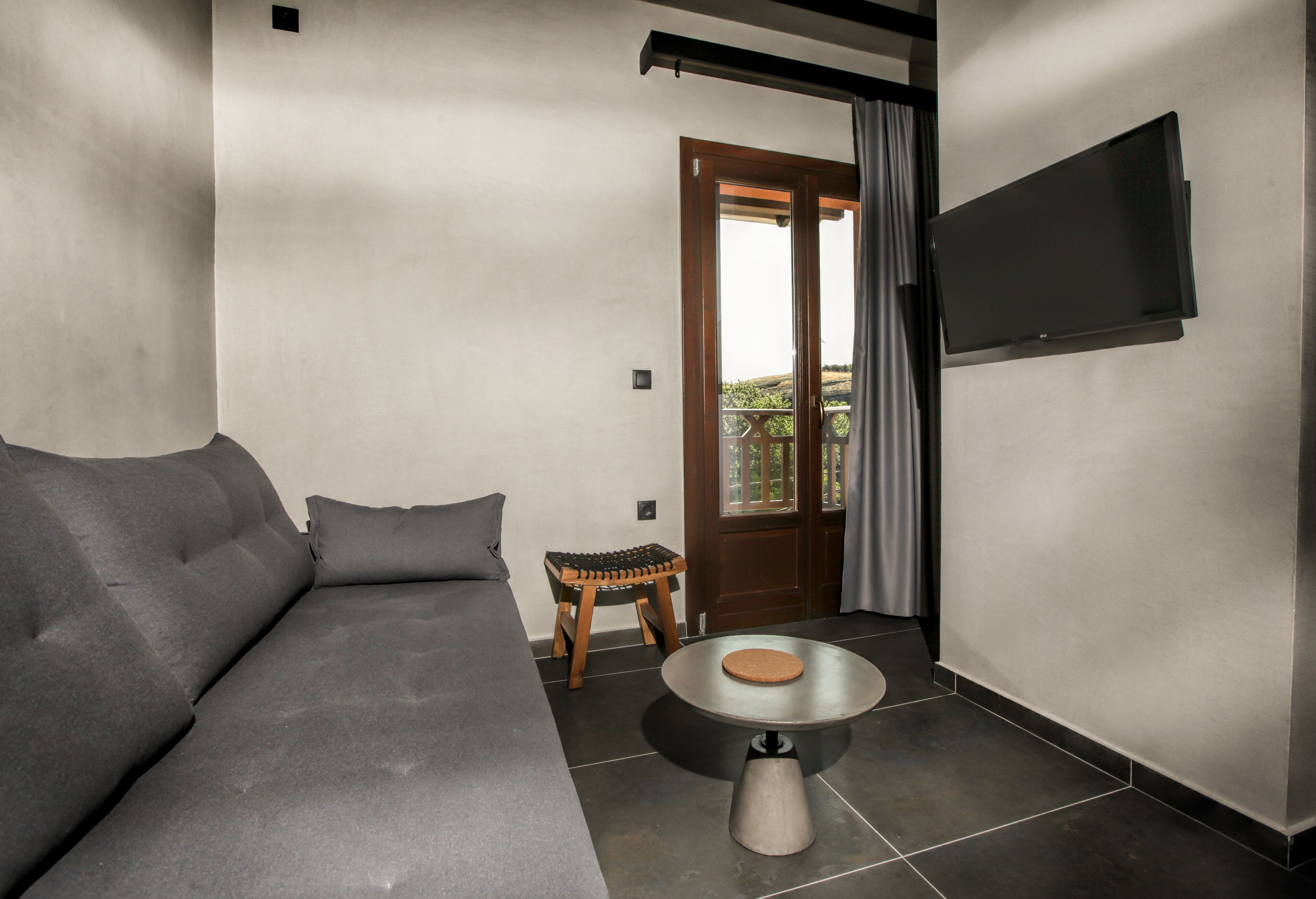 A modern living room with a grey sofa, a small wooden stool, a round metal table, and a TV mounted on the wall.
