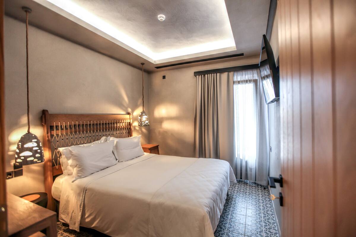 A hotel room with a large bed, a wooden headboard, a ceiling light, a TV mounted on the wall, and a window with curtains.
