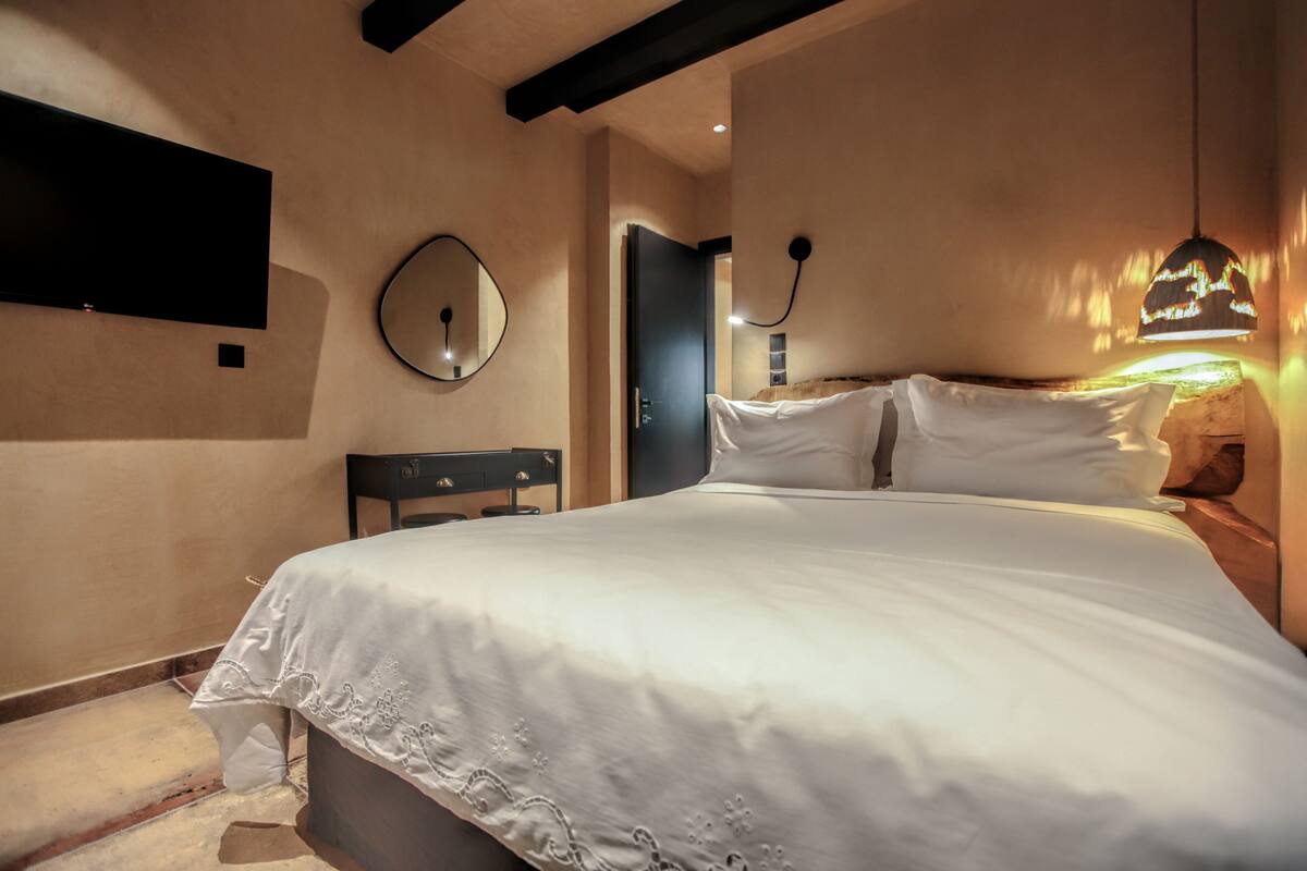 A neatly made bed with white linens, a round mirror on the wall, and a modern pendant light.