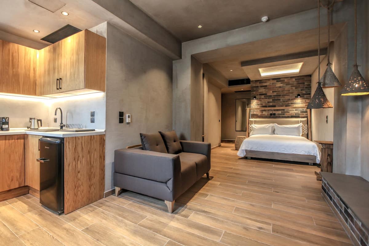 A modern kitchen with wooden cabinets and a built-in microwave. There is a grey sofa and a bed in the adjacent room.