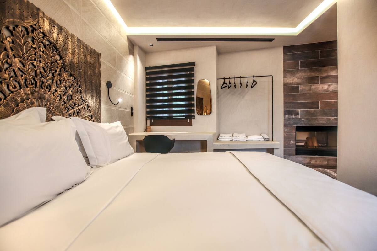 A modern hotel room with a large bed, a desk, a mirror, and a fireplace.