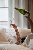 Una persona sosteniendo una botella de vino y una copa de vino en una habitación con ropa de cama blanca y una ventana.