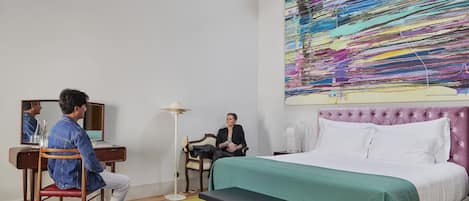 Een hotelkamer met een bed, een bureau, een stoel, een spiegel en een kleurrijk abstract schilderij aan de muur.