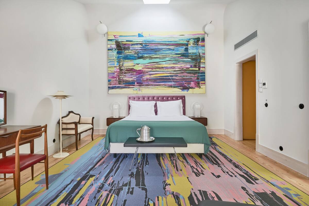 Habitación de hotel con un cuadro abstracto grande, una cama con colcha verde, una mesita de noche con jarrón plateado, una silla de madera y una lámpara de pie.