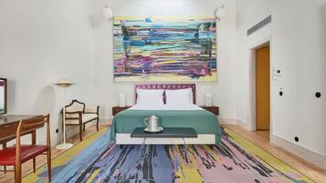 A hotel room with a large abstract painting, a bed with a green bedspread, a bedside table with a silver pitcher, a wooden chair, and a floor lamp.