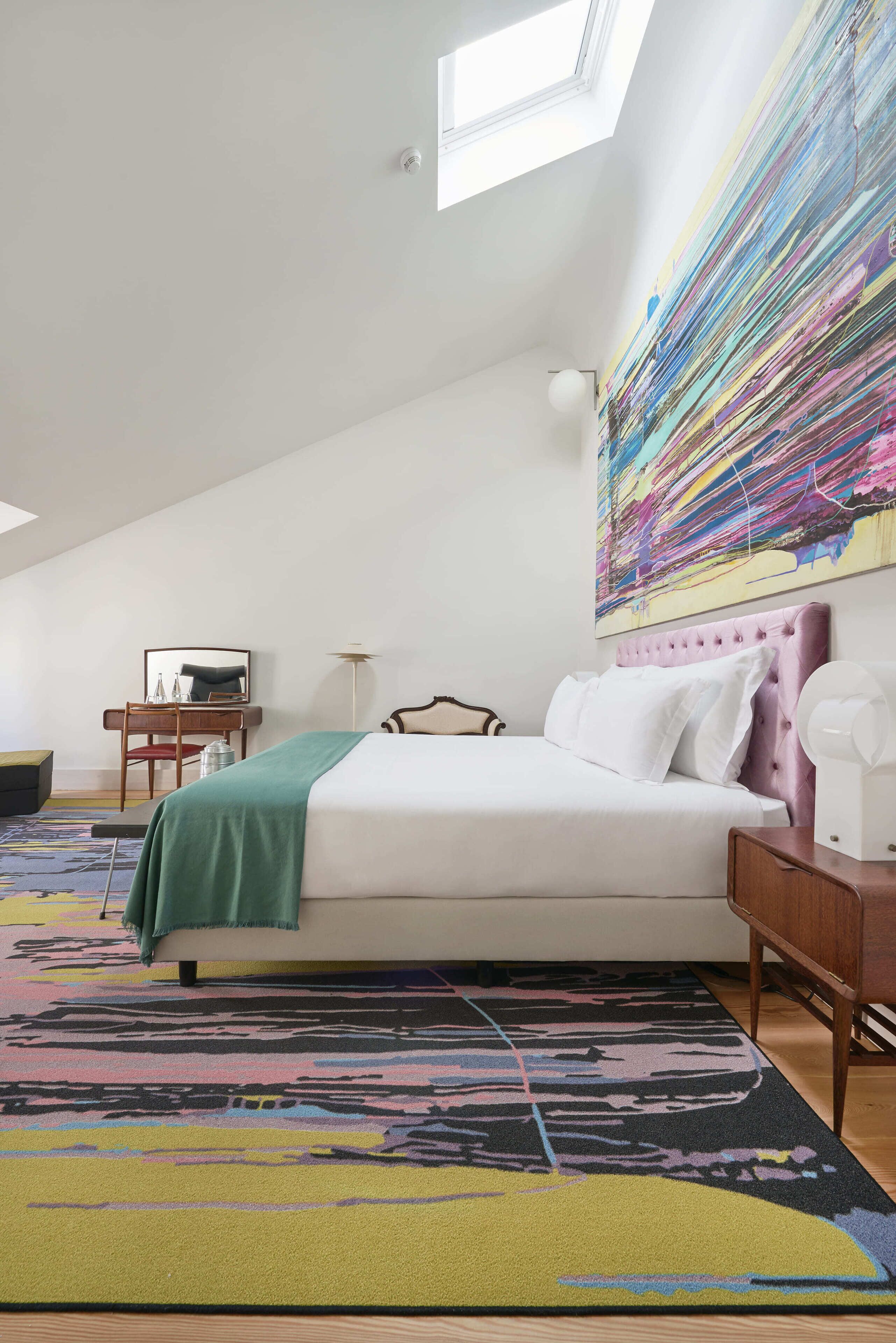 A bedroom with a bed, a desk, a chair, a large abstract painting, and a skylight.