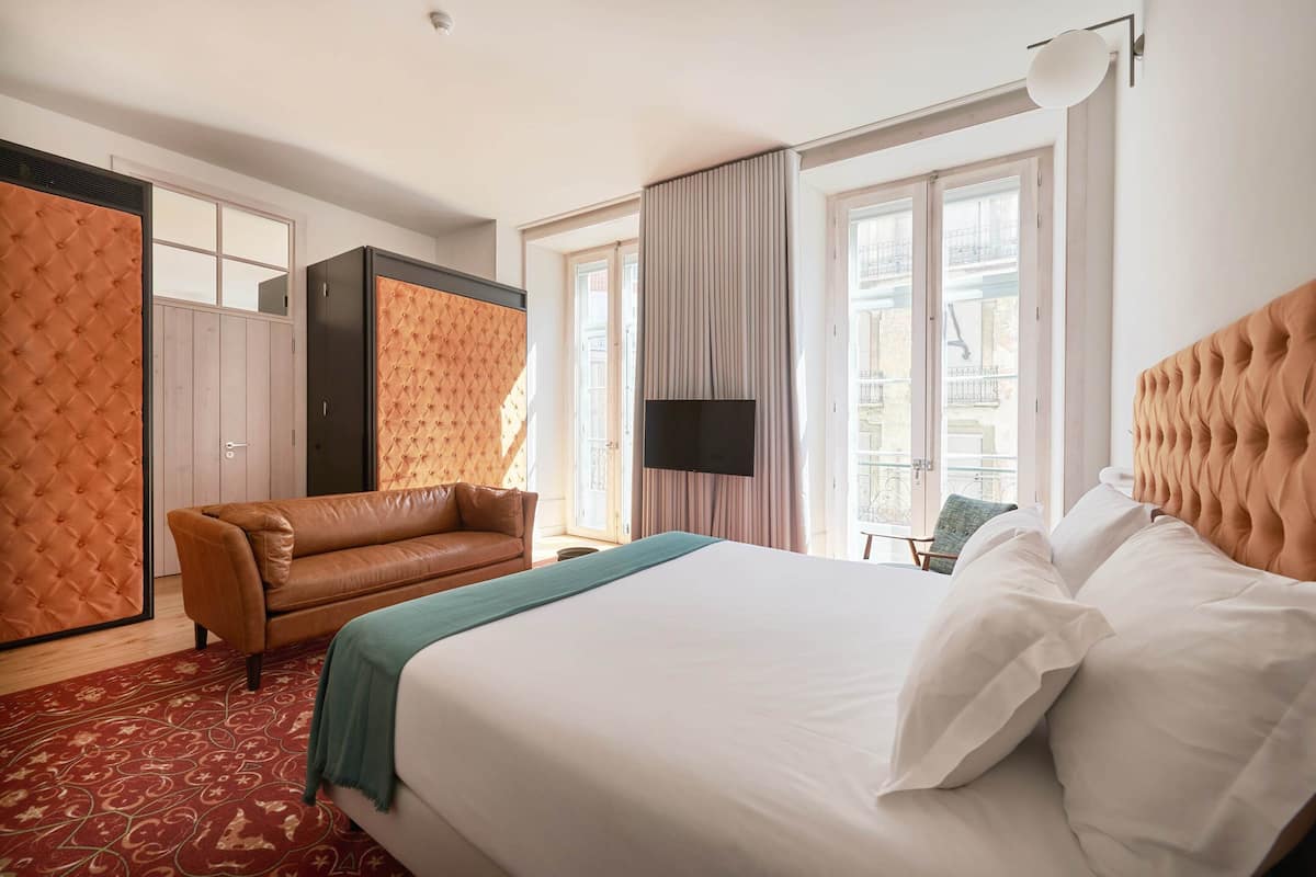 Habitación de hotel con una cama grande, un sofá de cuero marrón, un televisor y una ventana con cortinas.