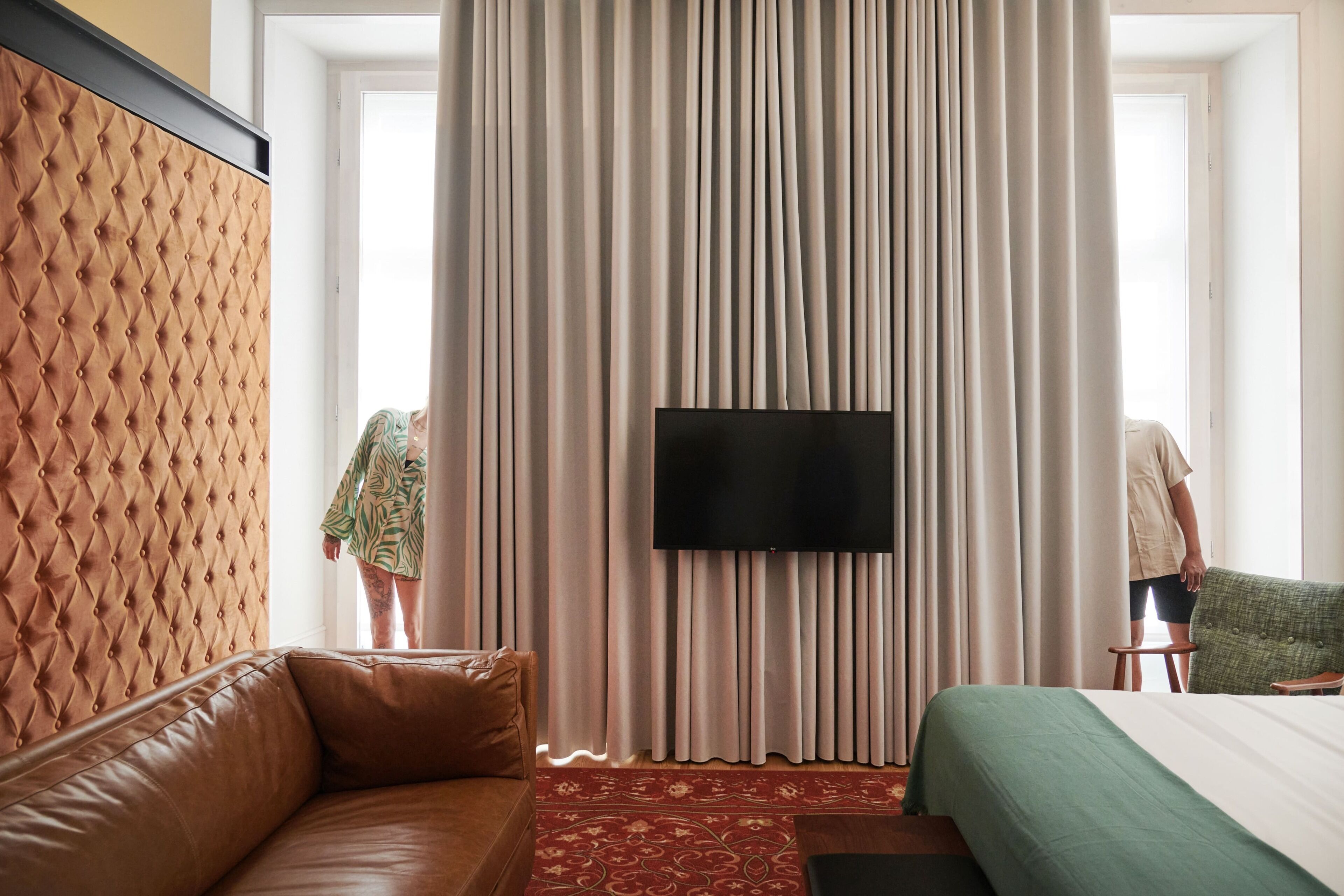 Une chambre d’hôtel avec une tête de lit capitonnée, un grand téléviseur à écran plat, une couvre-lit verte et un tapis rouge à motifs.